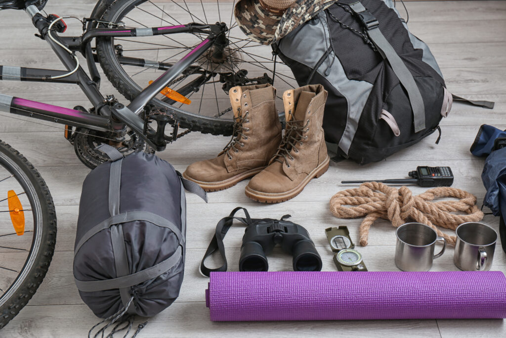 A picture of some outdoor equipment. A bike, a pair of hiking books, a sleeping bag, a rope and two cups. 