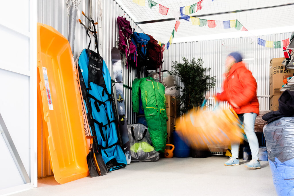 Lady in a red jacket lifting a big yellow bag. She is inside a storage unit fillet with outdoor activity equipment. 